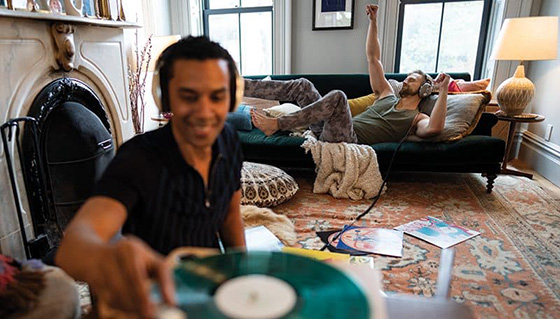 1 man being a DJ on a record player and another man laying on the couch listening to the music with headphones.